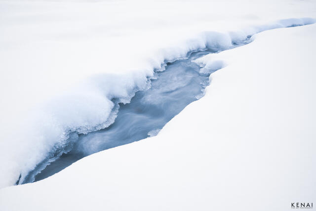 A stream cuts through deep snow in the winter of Alaska, near Black Rapids.