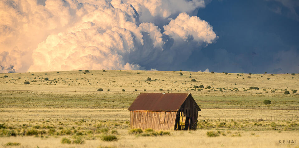 A weathered old barn stands on the plains with a powerful thunderstorm in the background.